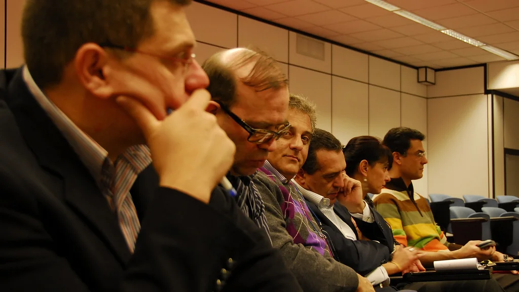 Persone sedute in una sala durante una riunione di lavoro, alcune con quaderni e appunti